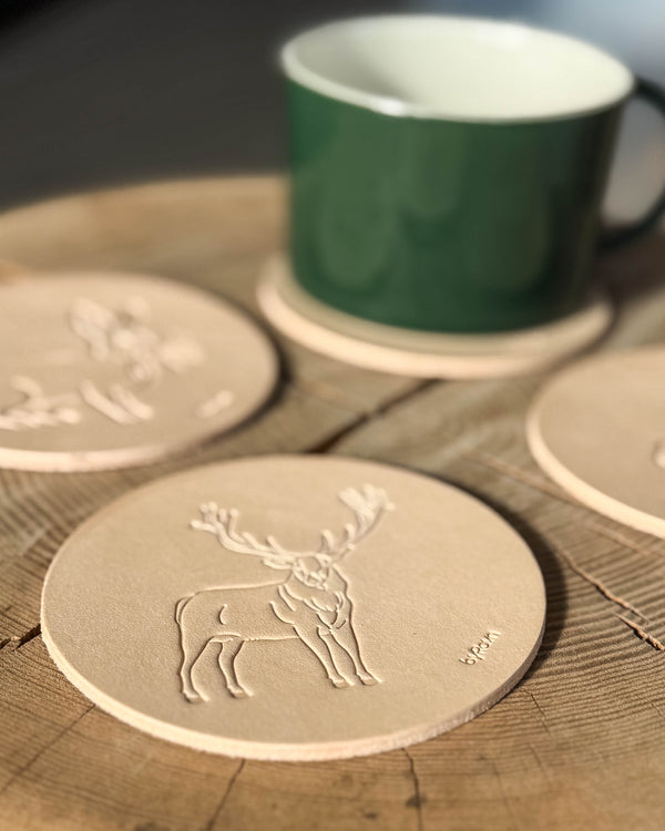 MOKKA jagt bordskåner i naturfarvet kernelæder præget med kronhjort på træbord, flere coasters og grøn kop bagved