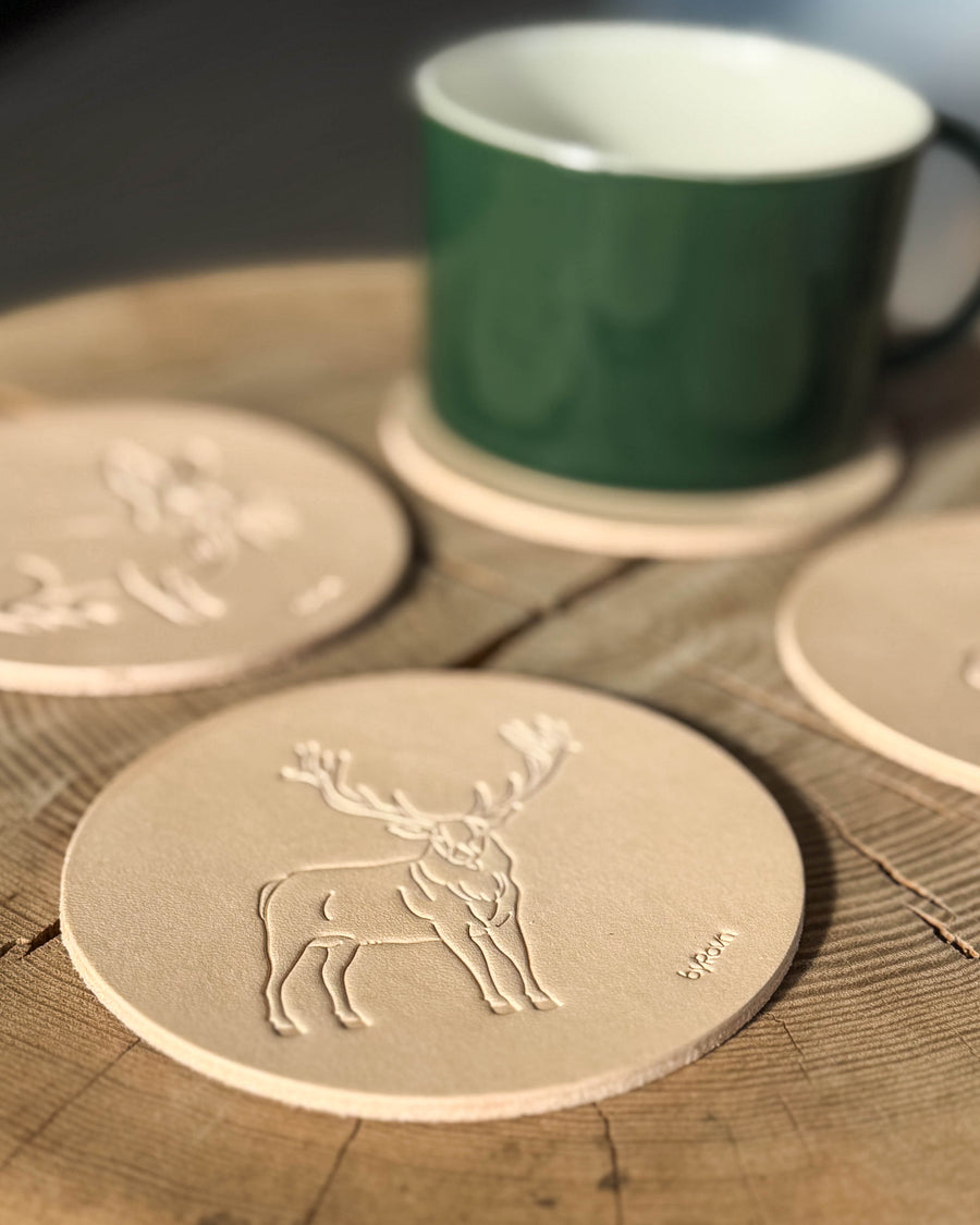 MOKKA jagt bordskåner i naturfarvet kernelæder præget med kronhjort på træbord, flere coasters og grøn kop bagved