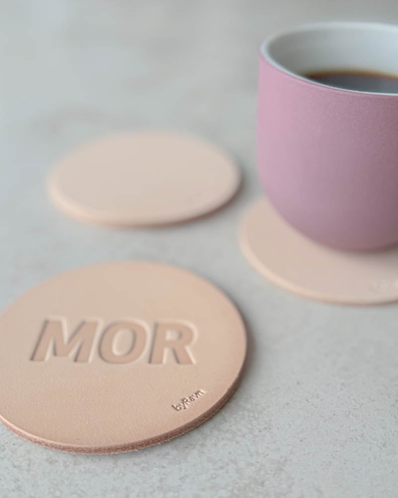 MOKKA bordskåner i naturfarvet kernelæder præget "Mor" i store bogstaver, rosa kaffekop på coaster bagved
