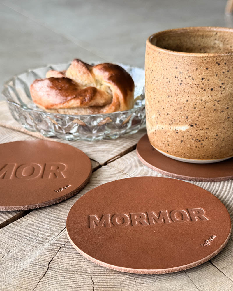 MOKKA bordskånere i cognac læder præget "Mor" og "Mormor" på træbord med kaffekop og kanelsnegl i glasskål