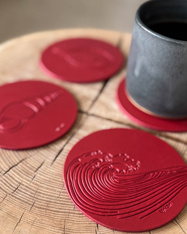 MOKKA sommerhus bordskånere i rødt læder præget med bølge og konkylie på træskive ved grå kaffekop på coaster