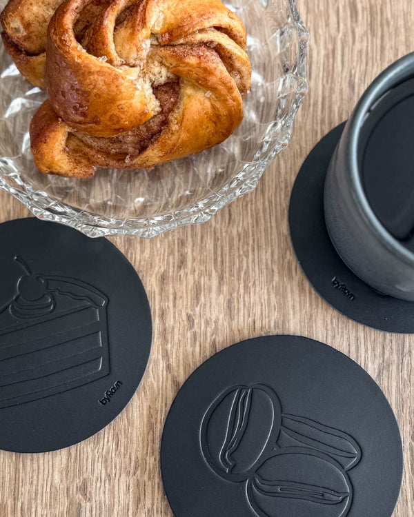 MOKKA kaffe & kage bordskånere i sort læder præget med lagkage og kaffebønner på træbord ved kanelsnegl og grå kop