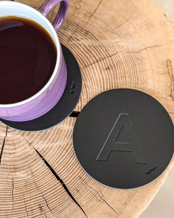 MOKKA bordskåner i sort læder præget med bogstavet "A" på træskive ved lilla kaffekop på coaster