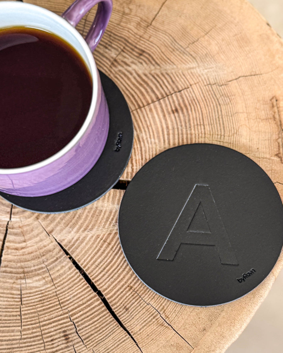 MOKKA bordskåner i sort læder præget med bogstavet "A" på træskive ved lilla kaffekop på coaster