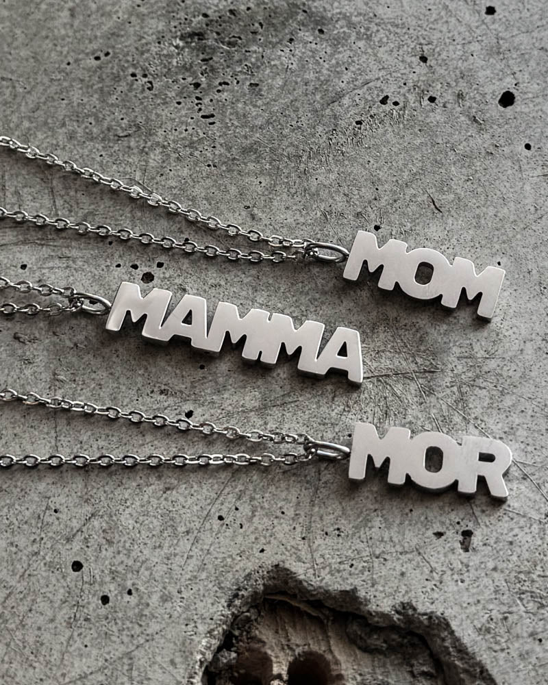 MOM halskæder i sølvfarvet rustfrit stål med bogstavvedhæng "MOM", "MAMMA" og "MOR", liggende på rå beton