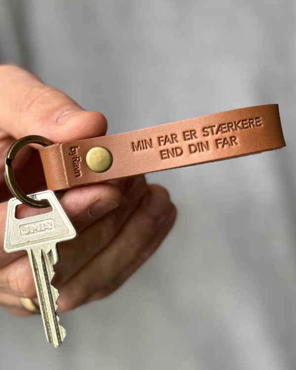 RAVN nøglering i cognac læder med graveringen "Min far er stærkere end din far", holdt i hånden med nøgle