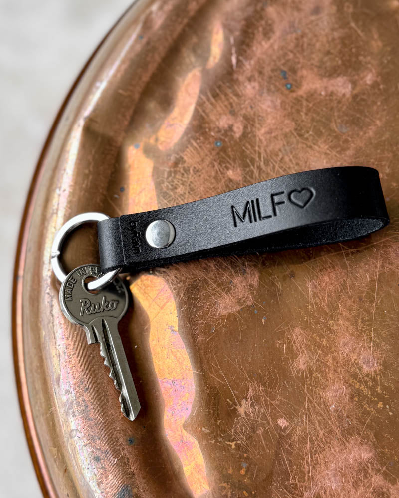 RAVN nøglering i mørkt læder med graveringen "MILF" ♡ og nøgle, liggende på patineret kobberbakke
