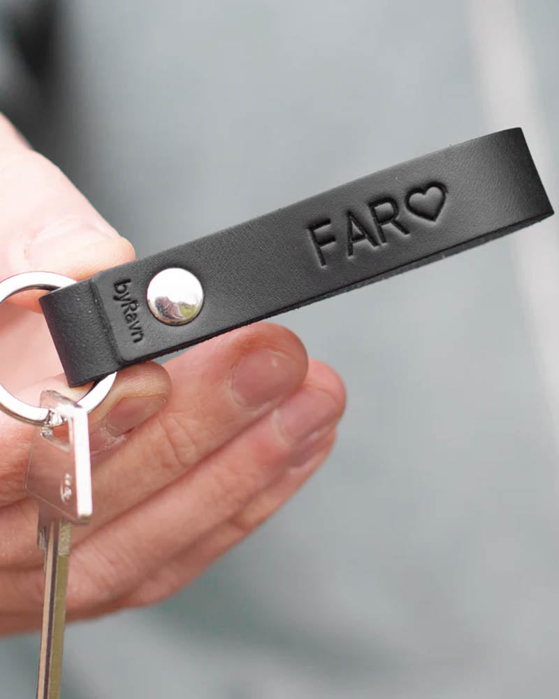 RAVN nøglering i sort læder med graveringen "Far" ♡ og nitte i stål, holdt i hånden med nøgle