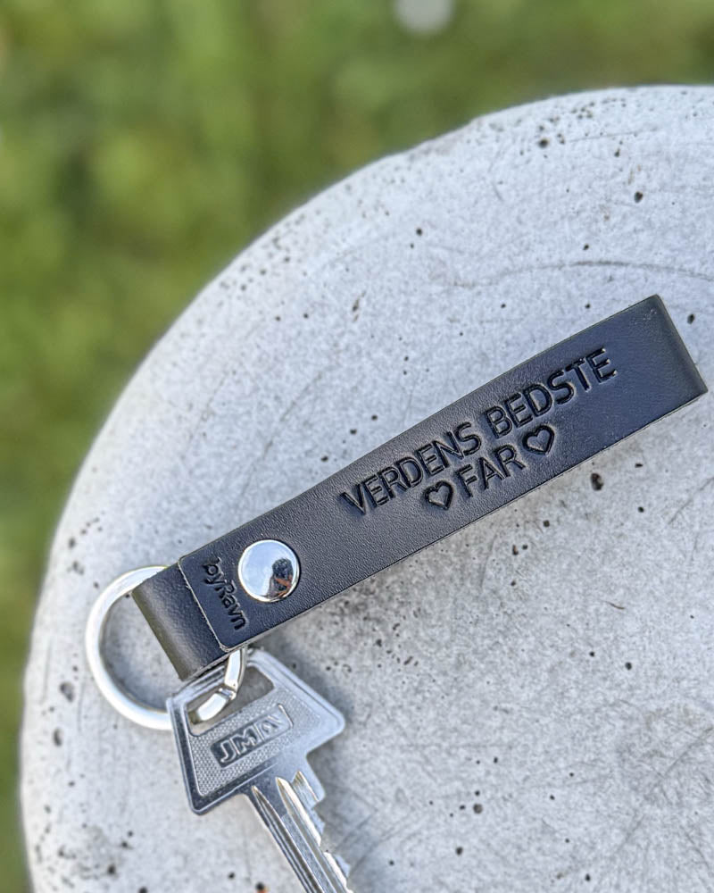 RAVN nøglering i sort læder med graveringen "Verdens bedste far" ♡ og nøgle, liggende på betonskive i græsset