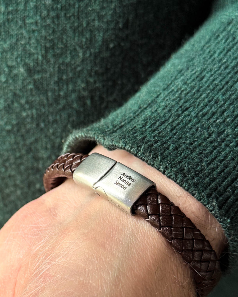 RAY læderarmbånd i mørkebrunt flettet læder med navnene "Anders, Nanna, Simon" graveret på stållåsen, båret til grøn strik