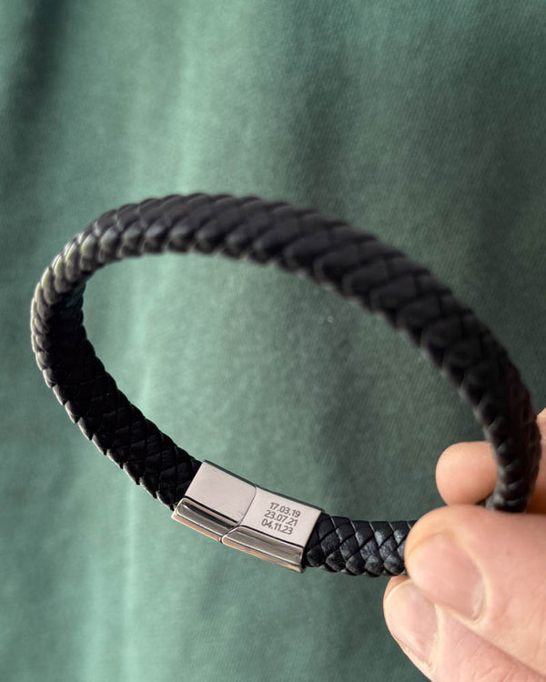 RAY læderarmbånd i sort flettet læder holdt i hånden, datoerne "17.03.19, 23.07.21, 04.11.23" graveret på låsen