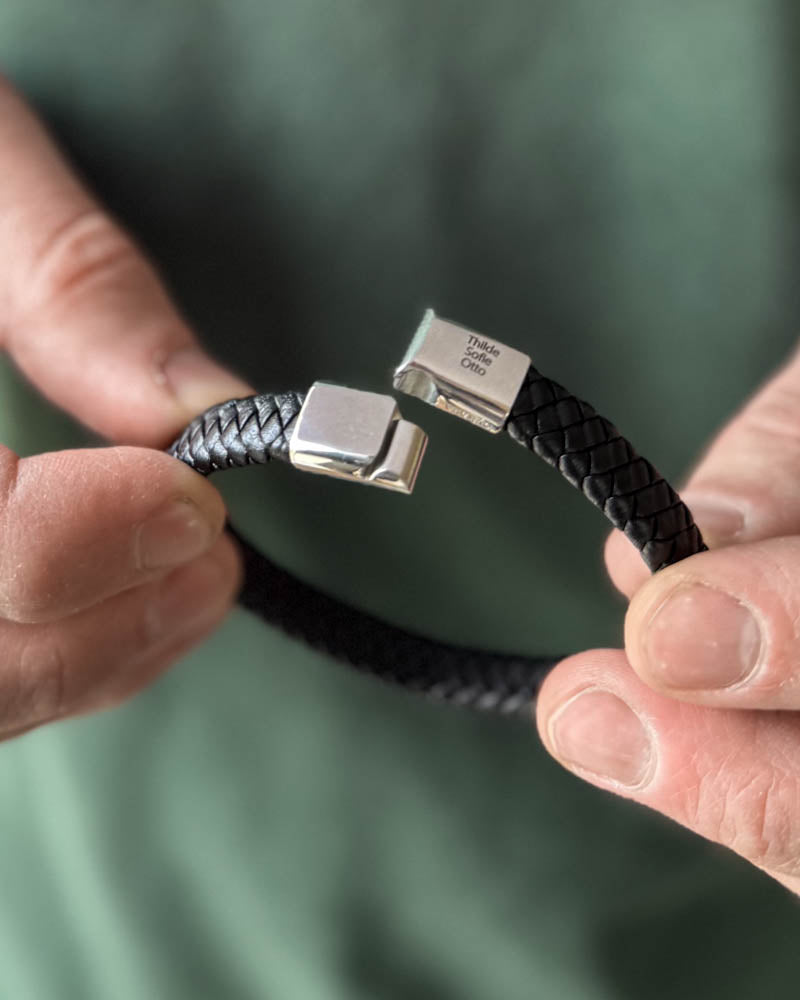 RAY læderarmbånd i sort flettet læder med åben magnetlås og navnene "Thilde, Sofie, Otto" graveret, holdt i hænderne