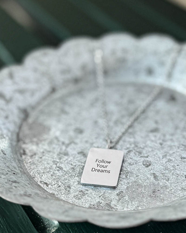 RAZ halskæde i sølvfarvet rustfrit stål med graveringen "Follow Your Dreams" på rektangulær plade, på hvidpatineret zinkfad