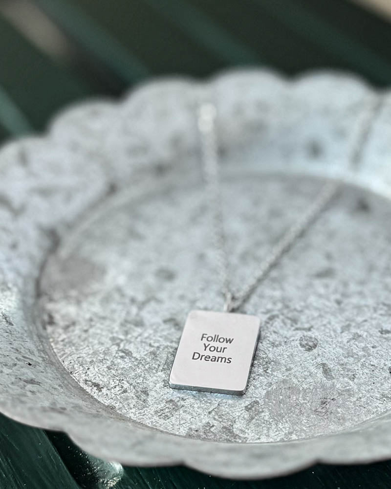 RAZ halskæde i sølvfarvet rustfrit stål med graveringen "Follow Your Dreams" på rektangulær plade, på hvidpatineret zinkfad
