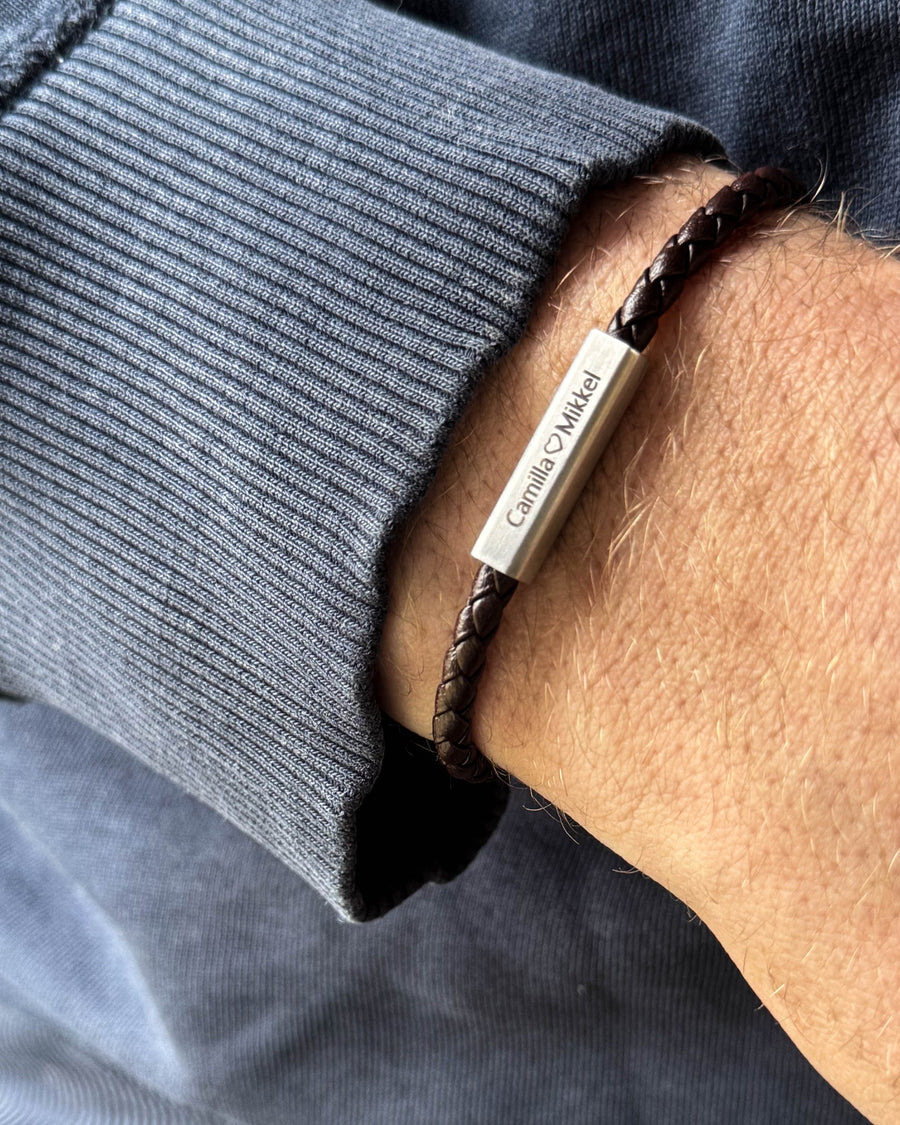 RIO læderarmbånd i mørkebrunt flettet læder med "Camilla ♡ Mikkel" graveret på stållåsen, båret om håndleddet
