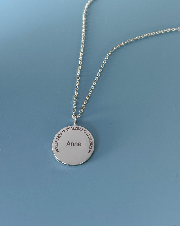 SIA CIRCLE navnehalskæde i sølvfarvet rustfrit stål med "Anne" og datoerne "21.03.2020 ♡ 09.11.2022 ♡ 17.06.2025" i cirkel, på blå bund