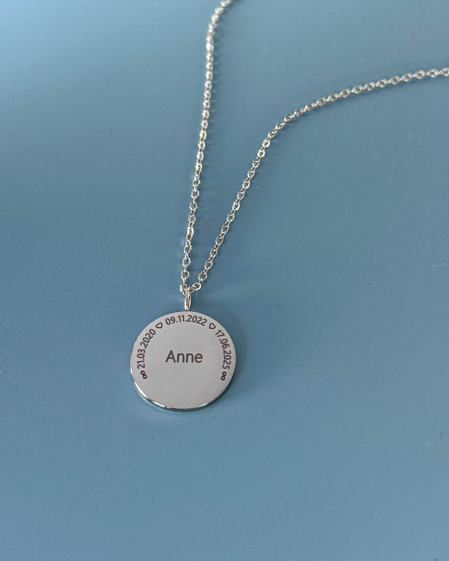 SIA CIRCLE navnehalskæde i sølvfarvet rustfrit stål med "Anne" og datoerne "21.03.2020 ♡ 09.11.2022 ♡ 17.06.2025" i cirkel, på blå bund