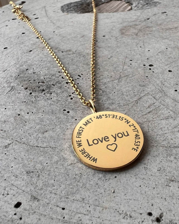 SIA CIRCLE halskæde i guldbelagt stål med "Love you" ♡ og "Where we first met" med koordinater graveret i cirkel, på beton