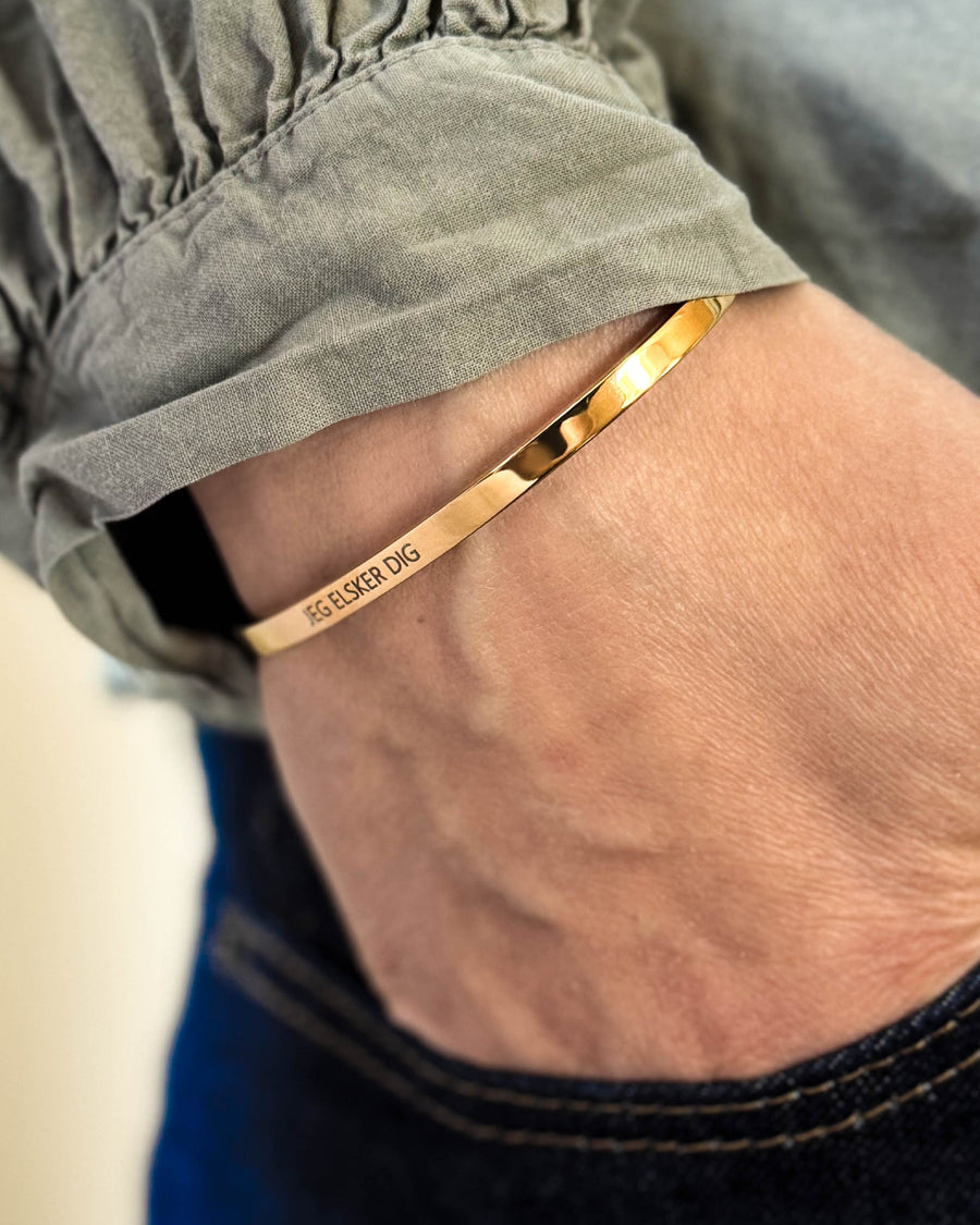 VENUS armbånd i guldbelagt stål med graveringen "Jeg elsker dig" om håndleddet, hånd i lommen på mørke jeans