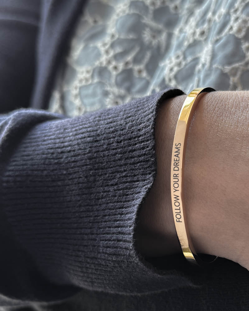 VENUS armbånd i guldbelagt stål med graveringen "Follow your dreams" i versaler, båret til mørkeblå strik og blondebluse