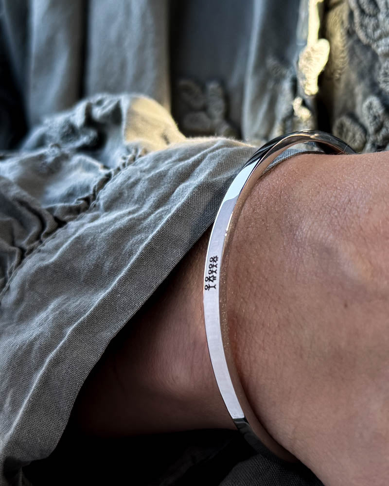 VENUS STICK armbånd i sølvfarvet rustfrit stål med tændstikfamilie graveret, båret til grå bluse med pufærmer